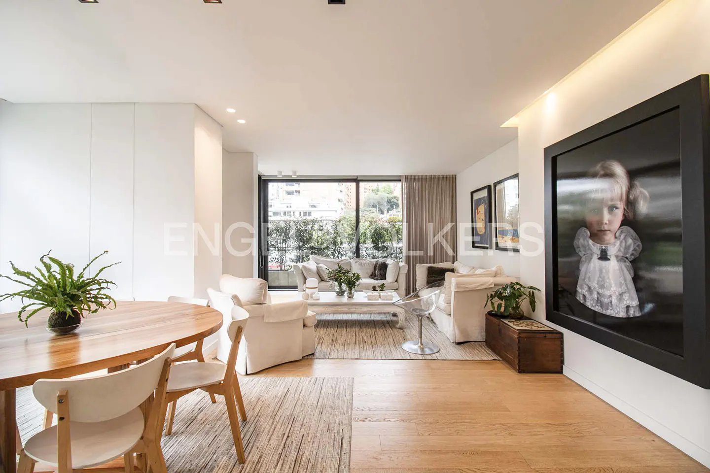 Bright living room with wood floors, white walls, and a large framed portrait. A round wood table with chairs sits near a white sofa and a balcony view.