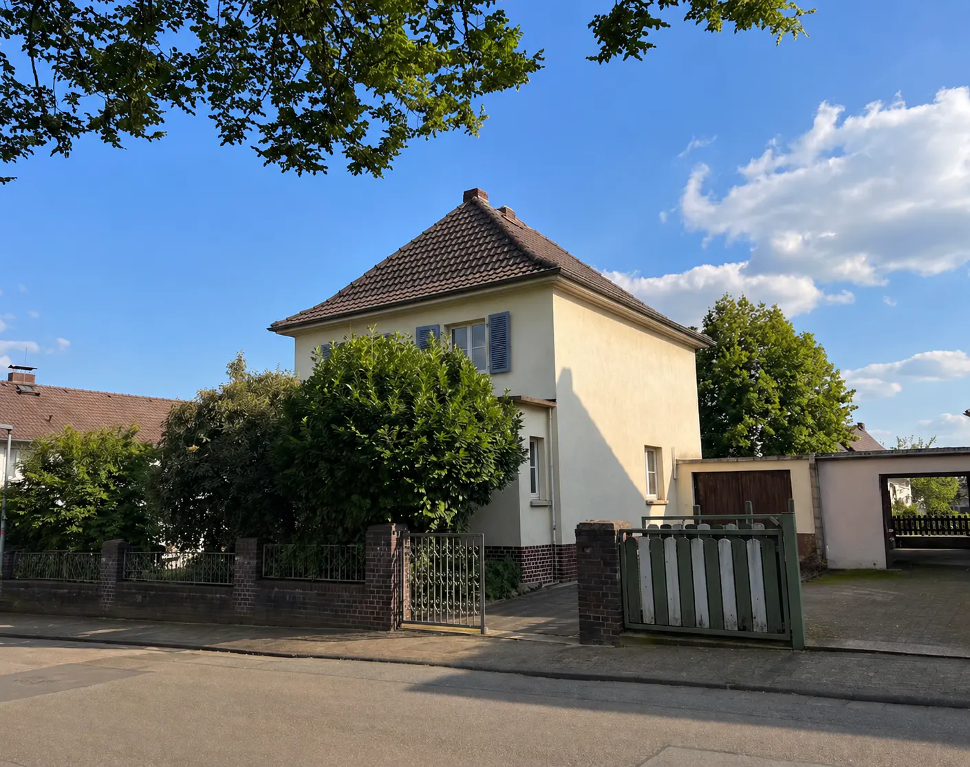 Two-story cream house with brown roof, blue shutters, and green trees under a blue sky. A brick and iron fence surrounds the property.