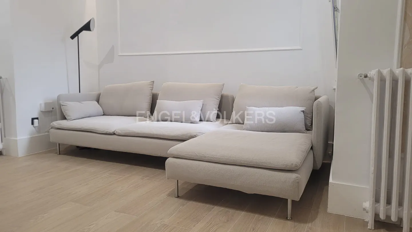 A modern living room features a beige sectional sofa with throw pillows, a black floor lamp, and a white radiator.