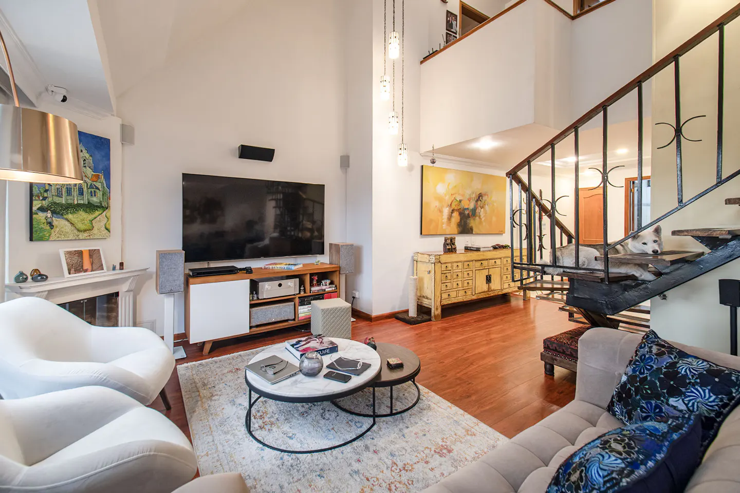 Bright living room with white walls, hardwood floors, and a staircase. A dog rests on the stairs. Modern furniture and artwork decorate the space.