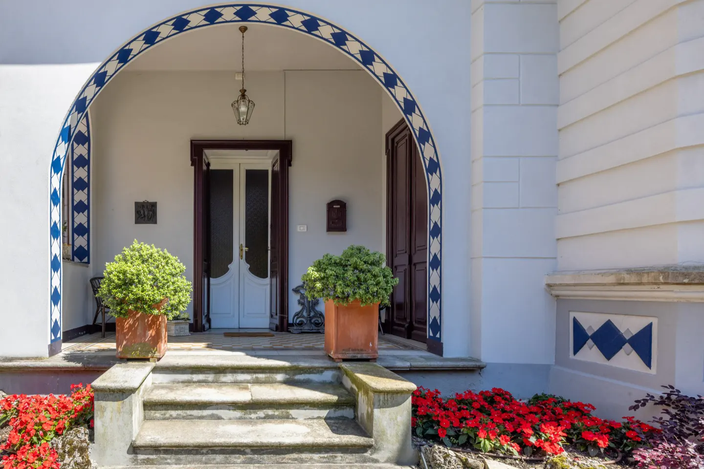Exterior view of a white house entrance with an arched doorway, potted plants, and red flowers.