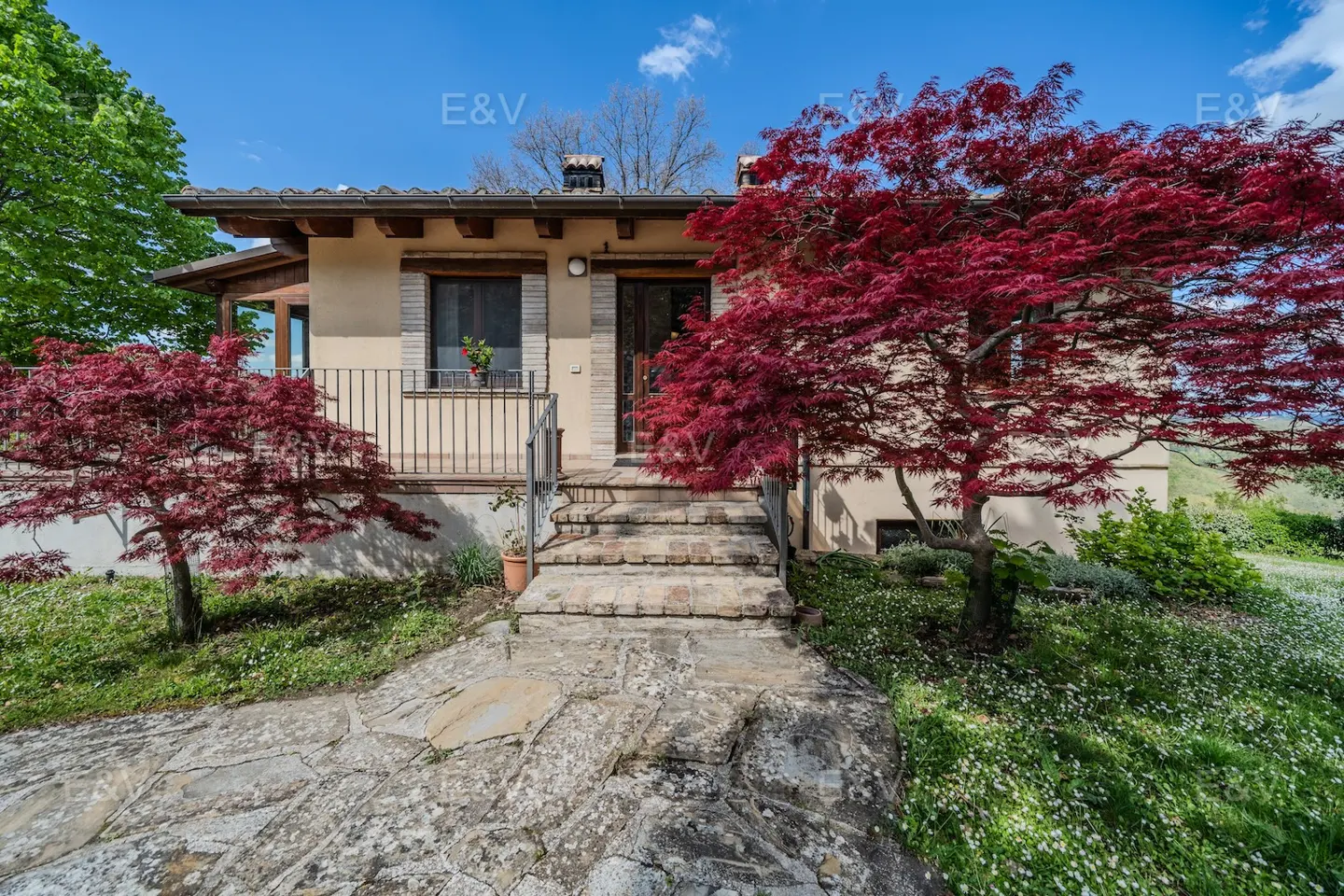 Exterior view of a beige house with a stone walkway, red trees, and a blue sky.