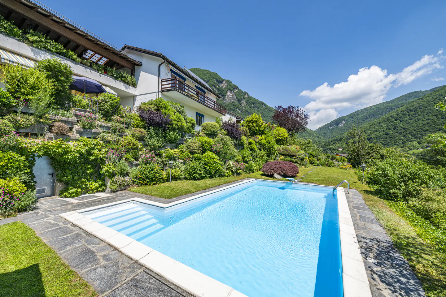 A bright blue swimming pool sits on a stone patio in front of a multi-level house surrounded by lush, terraced gardens.