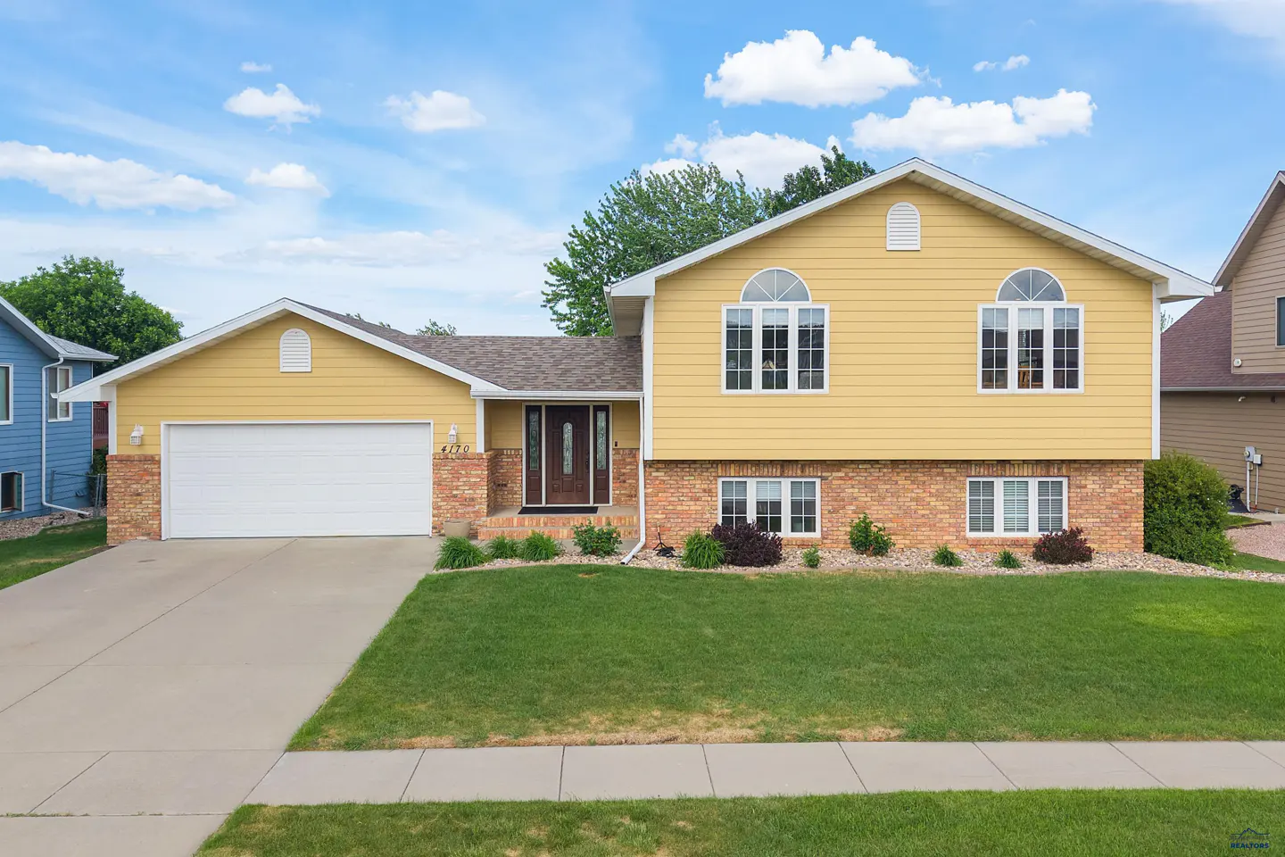 Yellow split-level home with a brick base, white garage door, and green lawn under a blue sky with scattered clouds.