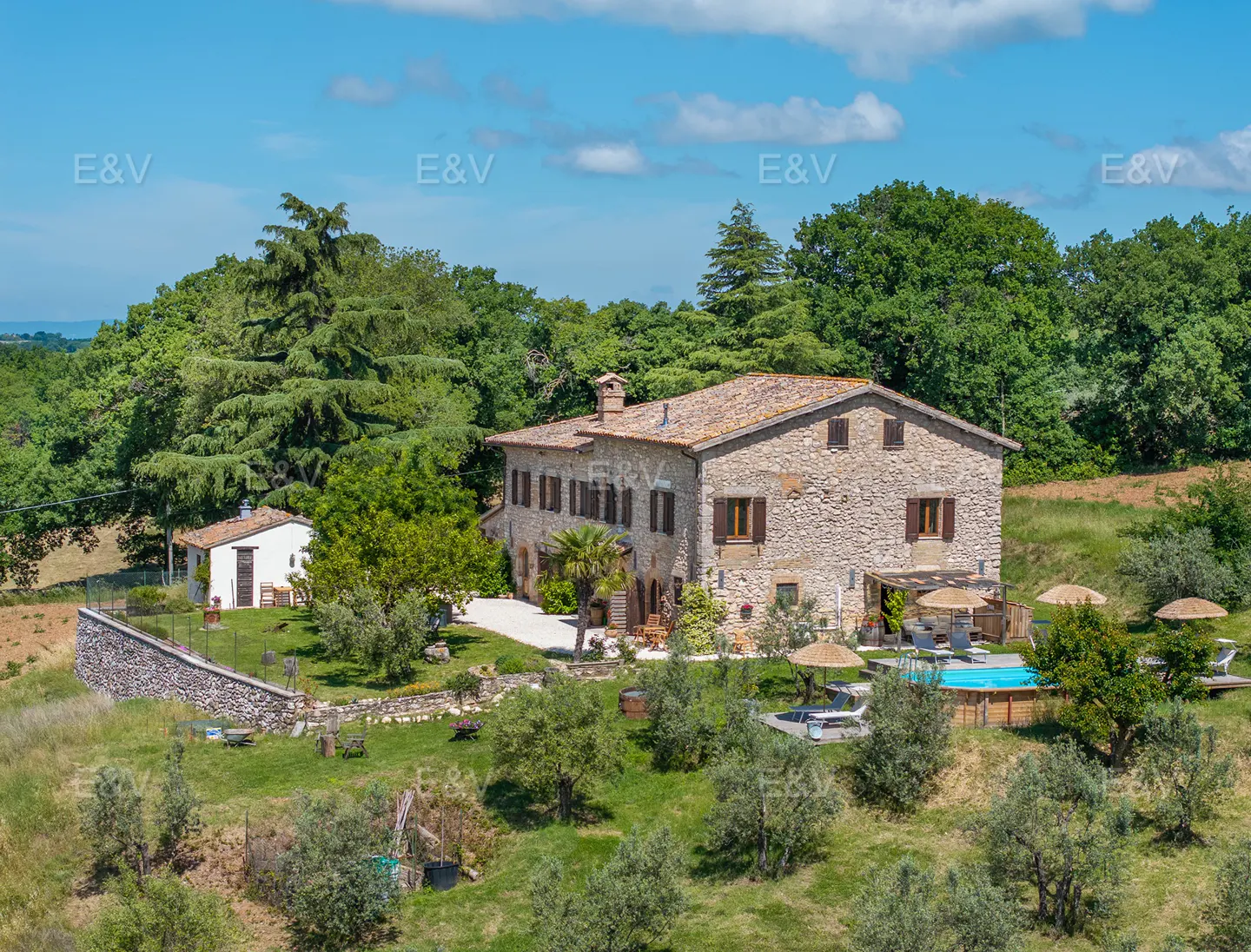 A large stone farmhouse with a tiled roof, surrounded by lush trees, a swimming pool, and a small white guest house.