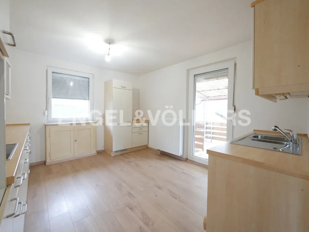 Bright kitchen with light wood cabinets, stainless steel sink, and wood floors. A window with blinds and a door to a balcony are visible.