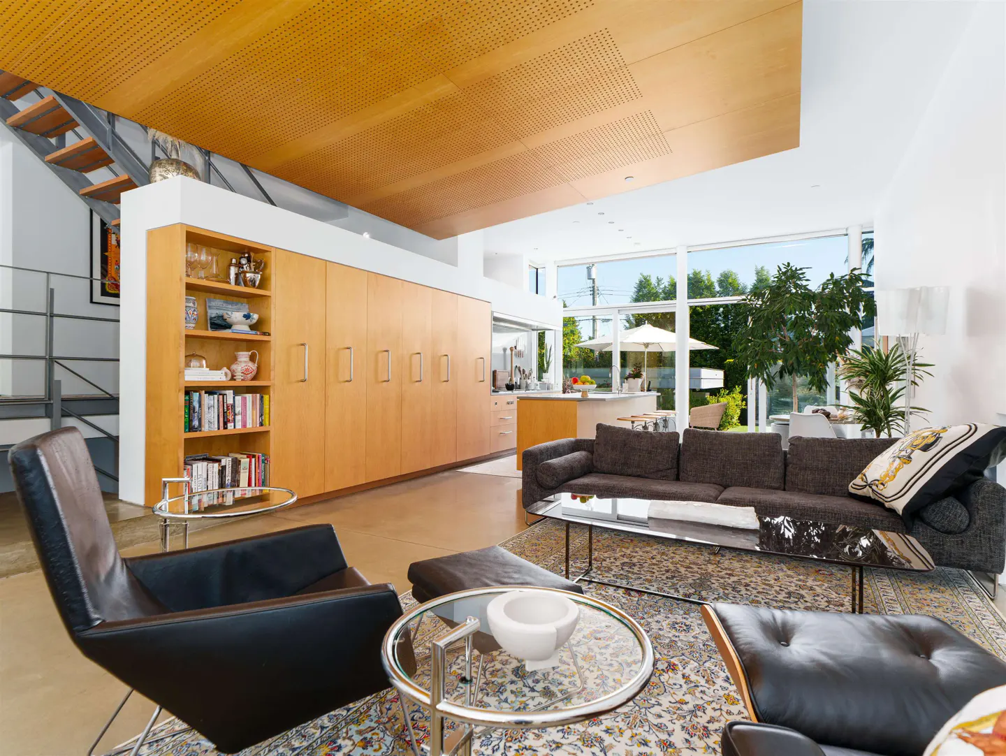 Modern living room with black leather chairs, brown sofa, and light wood cabinets. Large windows overlook a patio and garden.