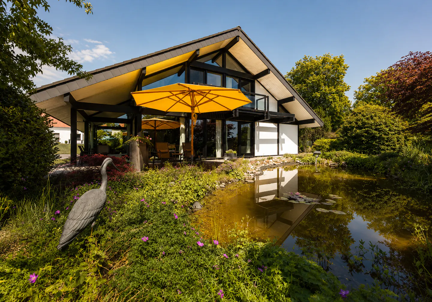 Modern A-frame house with a bright yellow umbrella on a patio, overlooking a garden pond with a heron statue.