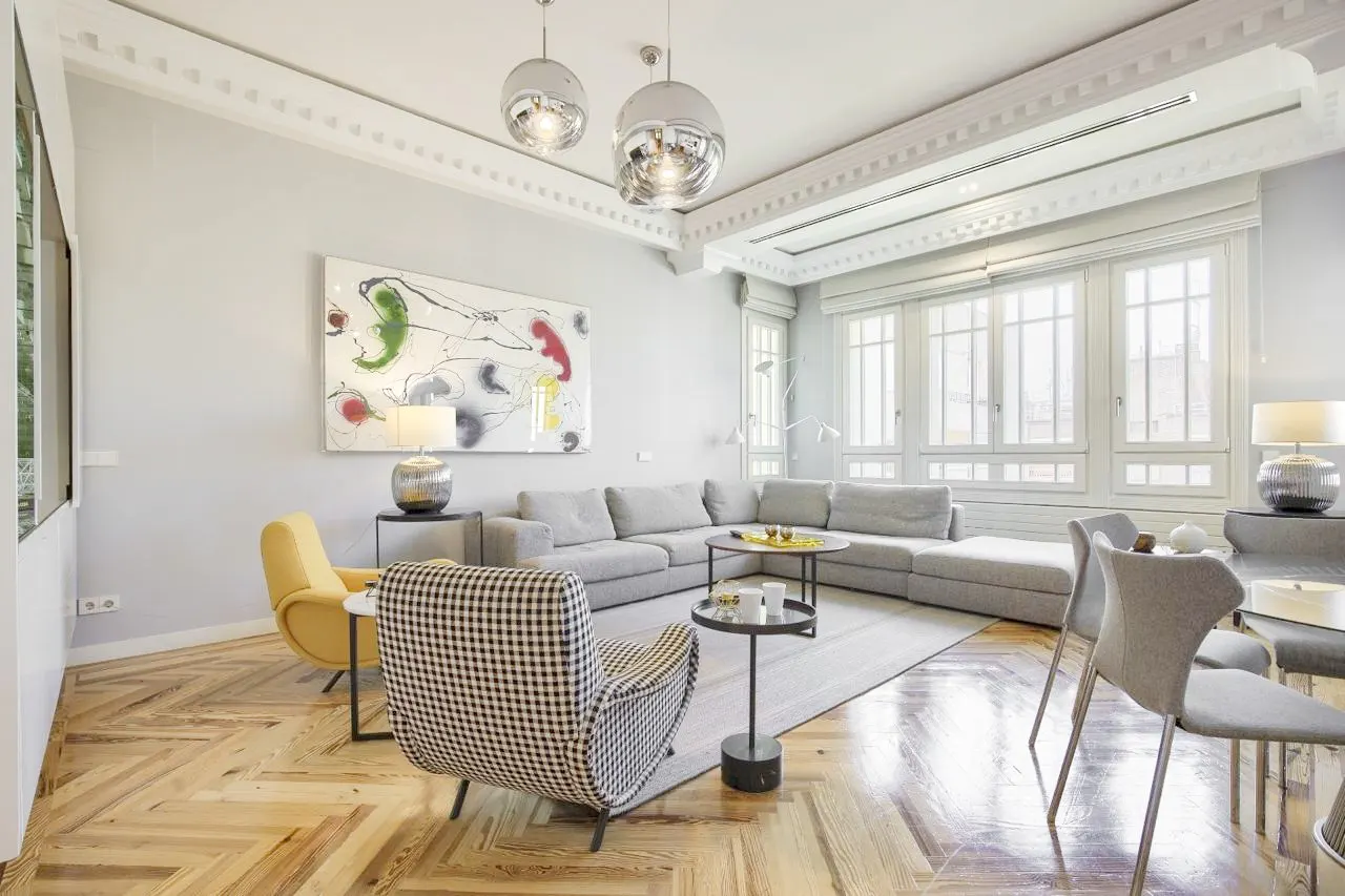 Bright living room with herringbone wood floors, a gray sectional, and a black and white checkered chair. Large windows and modern art.