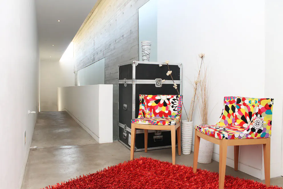 Hallway with concrete walls, a red rug, and two chairs with colorful floral patterns. A black trunk sits against the wall.