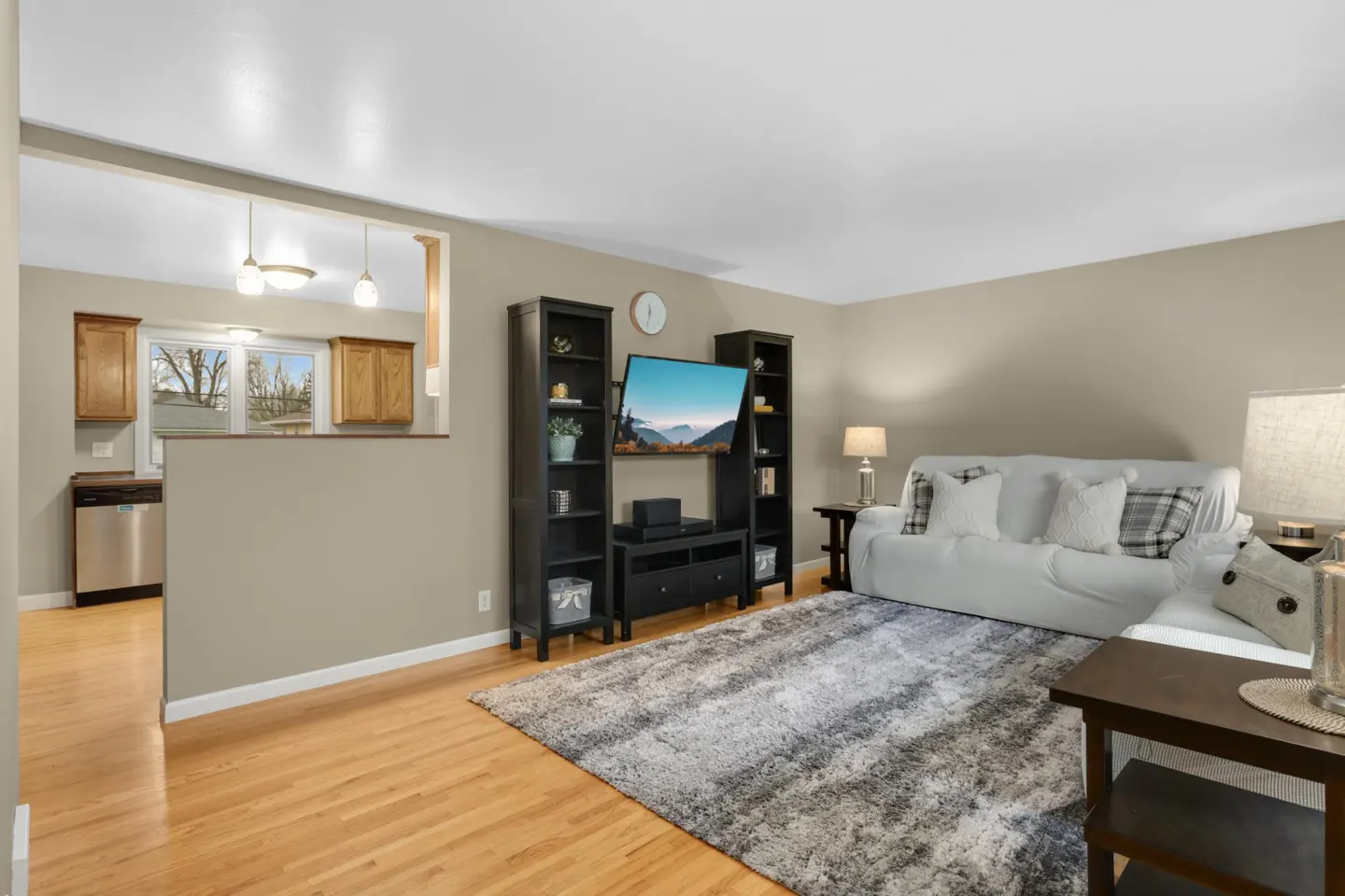 Living room with hardwood floors, a gray rug, and a white sofa. A black entertainment center holds a TV. The kitchen is visible through a wall opening.