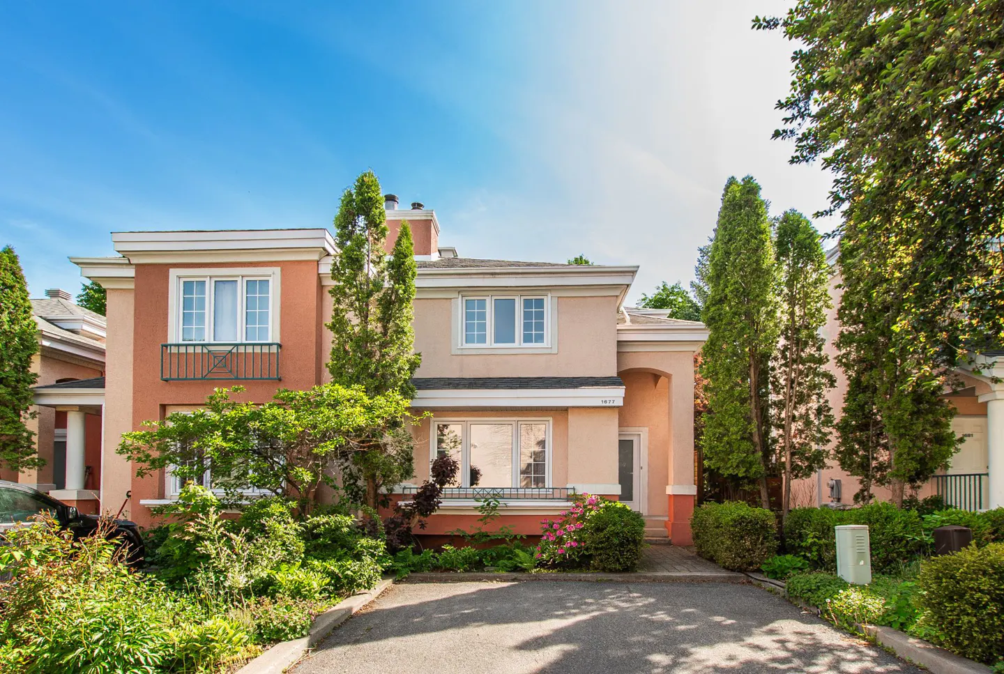 Two-story peach and tan house with a paved driveway, lush green landscaping, and tall evergreen trees under a blue sky.