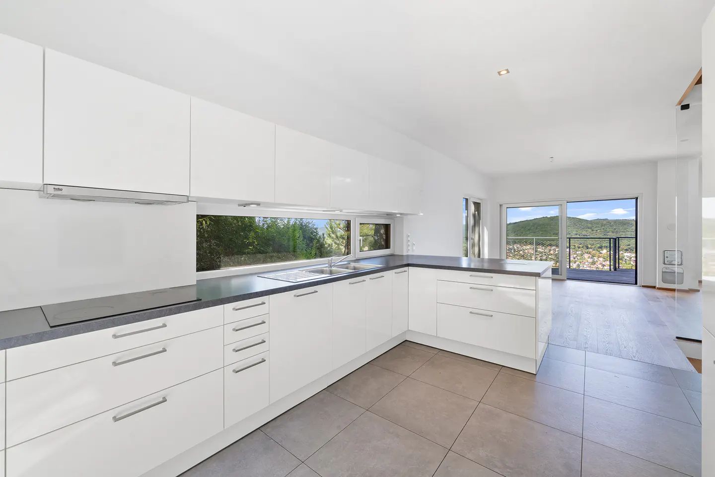 A modern kitchen with white cabinets, gray countertops, and gray tile flooring. A window overlooks a green landscape. Sliding glass doors lead to a balcony.