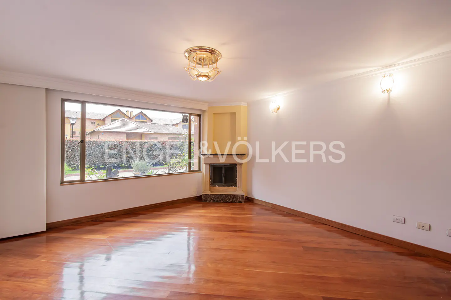 Empty living room with hardwood floors, a fireplace, and a large window showing a house. The walls are white, and there is a light fixture on the ceiling.