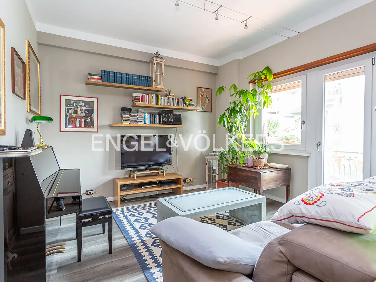 Living room with a black piano, blue patterned rug, and a gray sofa. Shelves with books and a TV are on the wall. A large plant sits near the window.