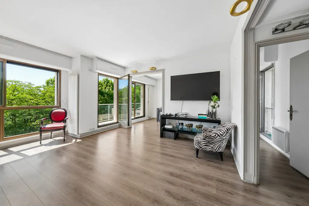 Bright living room with wood floors, white walls, and large windows overlooking green trees. A red chair sits near the window, and a zebra-print chair faces a TV.