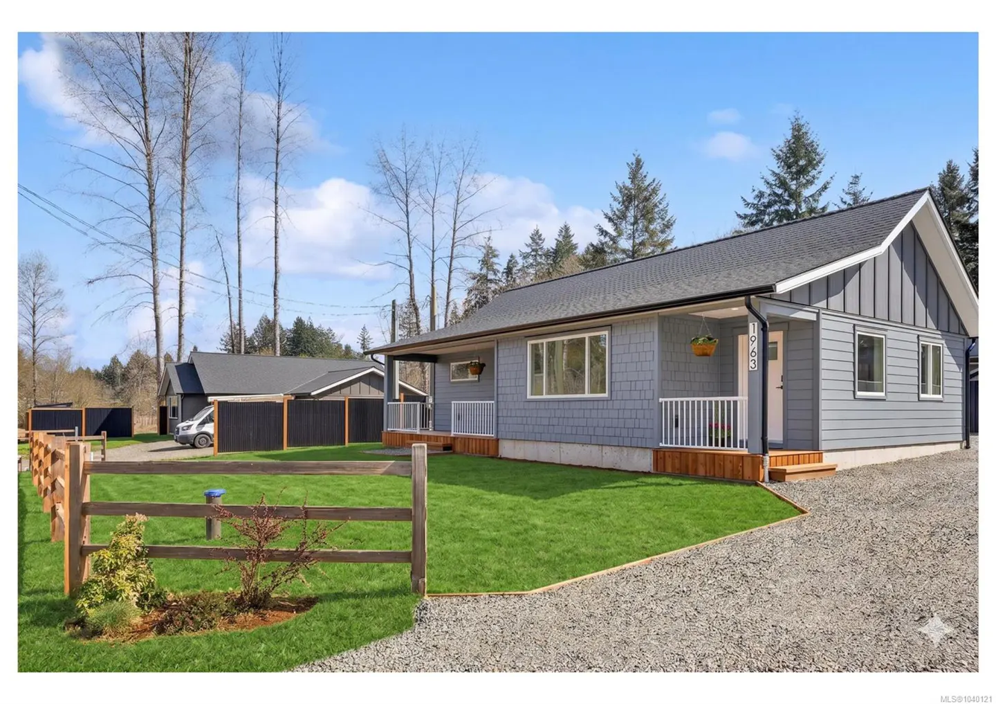 A grey, single-story house with a white porch and gravel driveway, set on a grassy lot with a wooden fence under a blue sky.