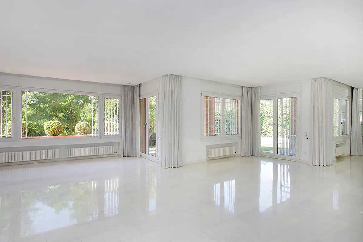 Bright, empty white room with large windows showing green trees and bushes outside. White curtains hang on either side of the windows.