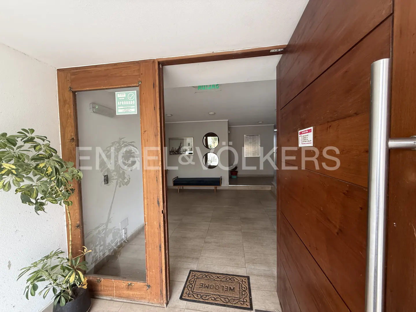 Open wooden door reveals a building lobby with tile floors, bench, and artwork. A potted plant sits near the door.