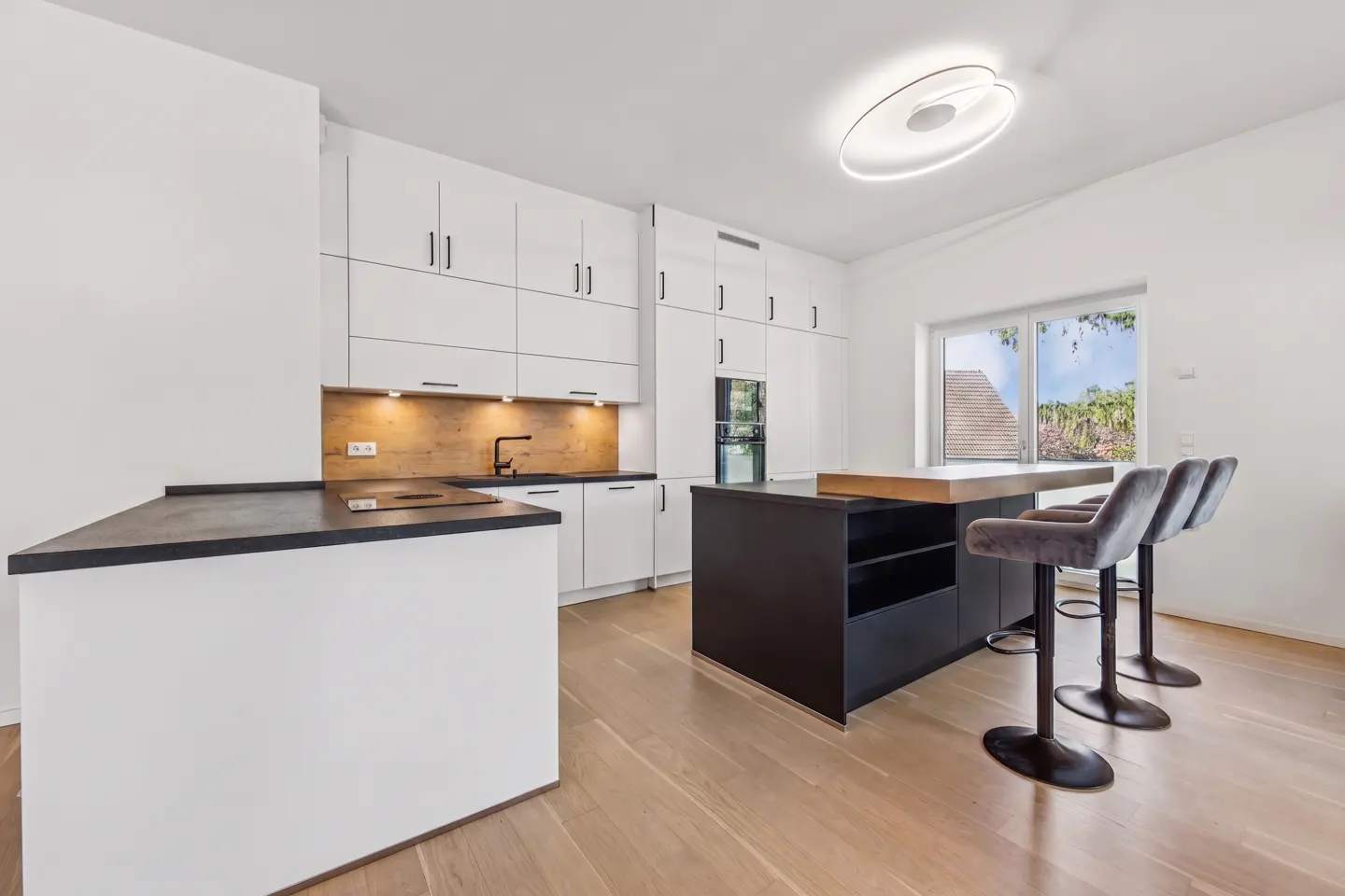Bright kitchen with white cabinets, black countertops, and a dark island with three gray velvet bar stools. Light wood floors and a window with a view.