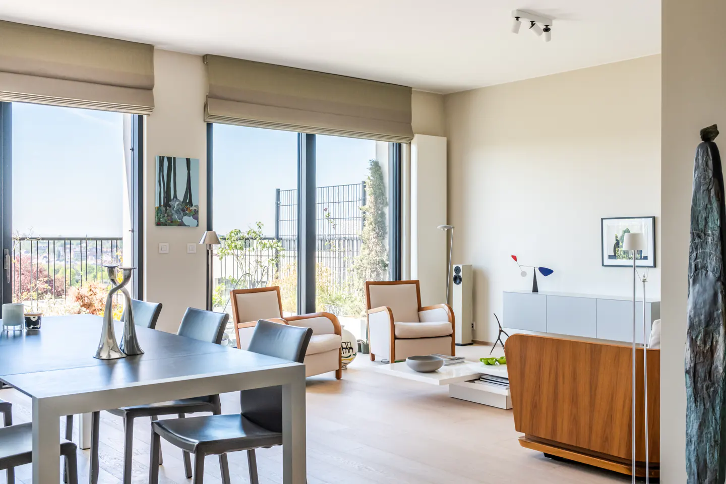 Bright, modern living room with a dining table, chairs, and large windows overlooking a cityscape. Neutral tones and minimalist decor.