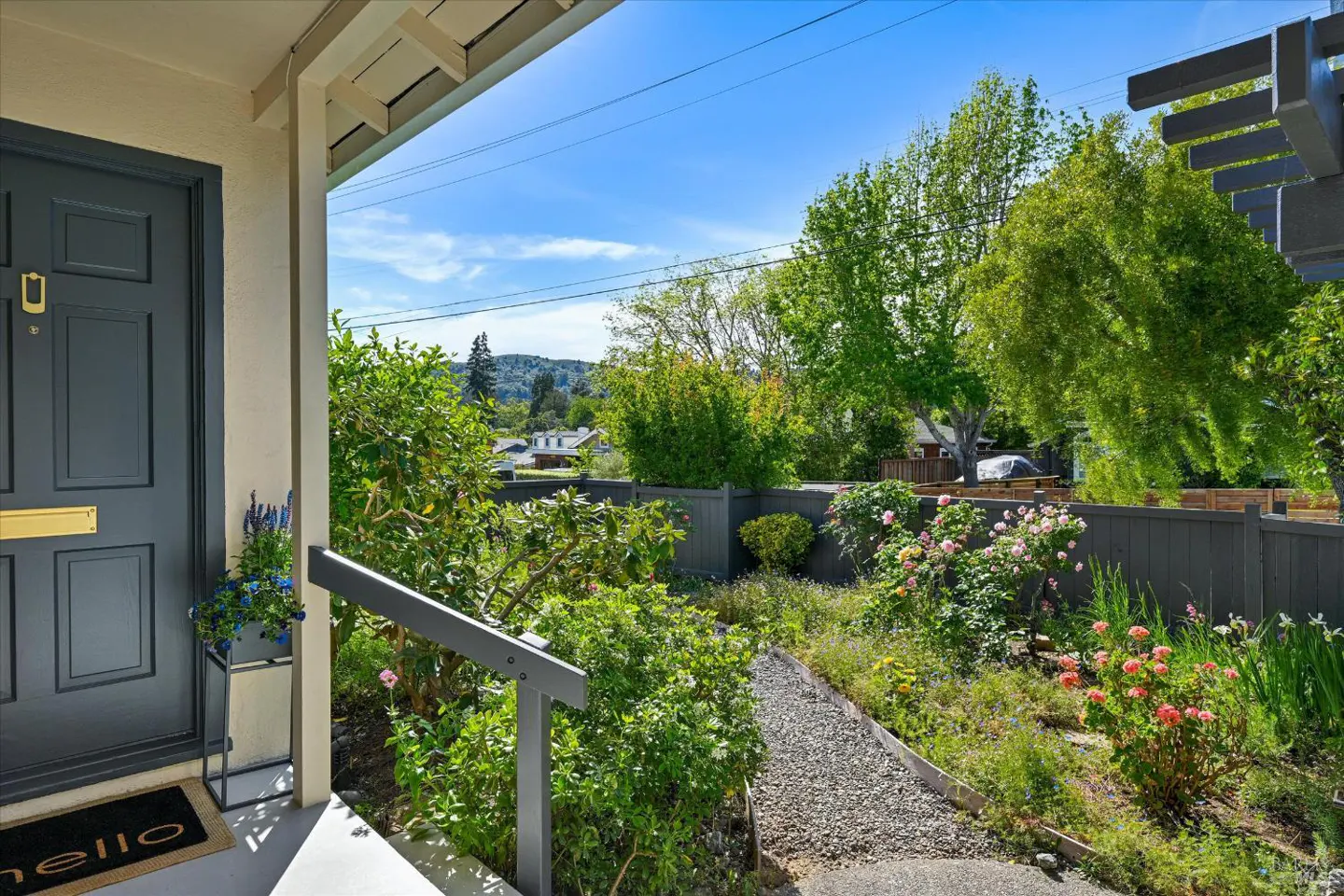 Front door view of a home with a gray door, gold hardware, and a "hello" doormat. A garden path leads to a gray fence with trees and a blue sky in the background.