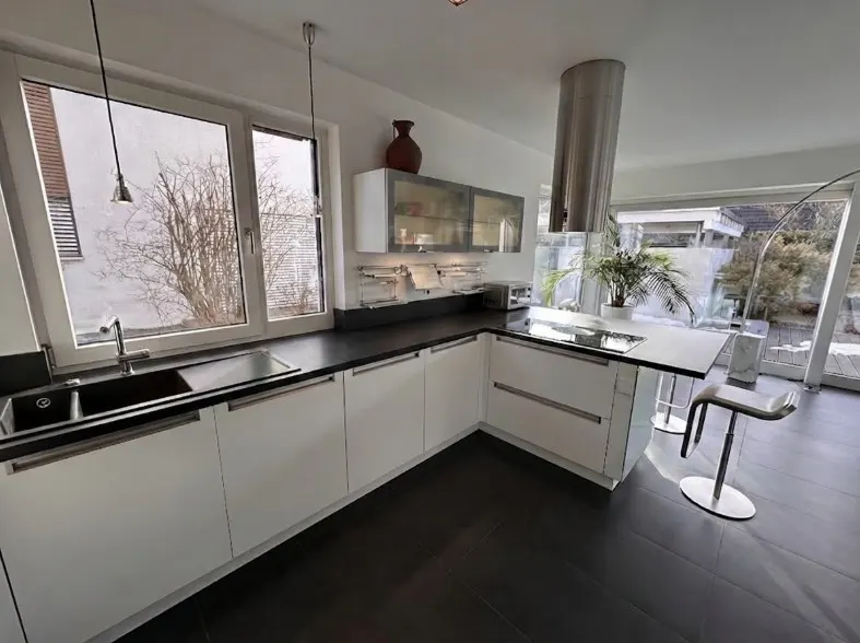 Bright kitchen with white cabinets, black countertops, and dark wood floors. Large windows offer a view of the outside.