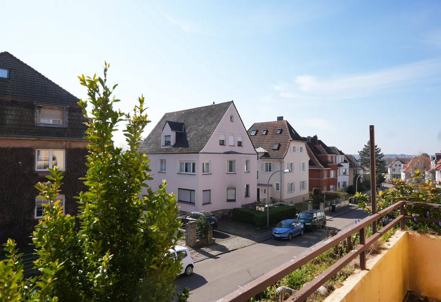 View from a balcony overlooking a street with houses and parked cars on a sunny day.