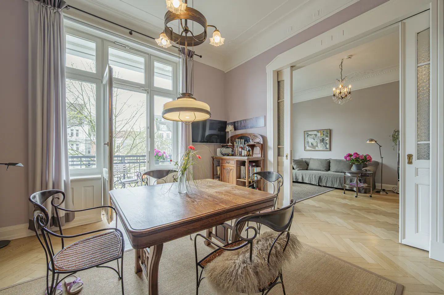 A dining room with a wooden table and chairs, a balcony with open doors, and a view into the living room.