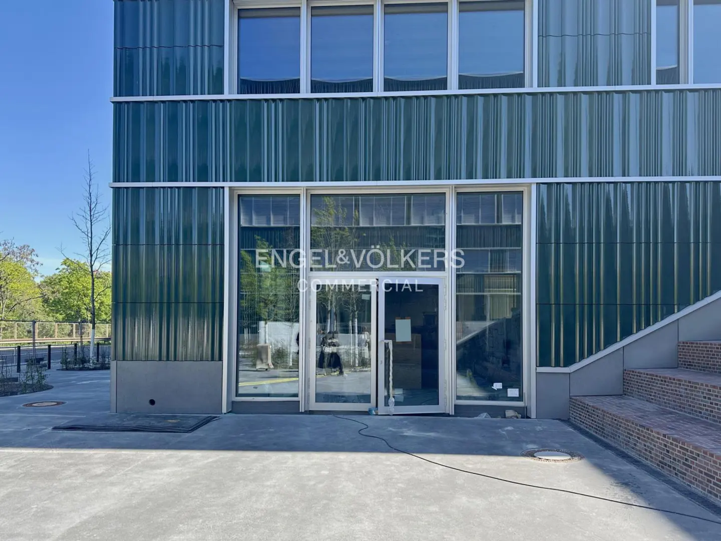 Exterior of a modern commercial building with green corrugated metal and glass doors with "Engel & Völkers Commercial" logo.