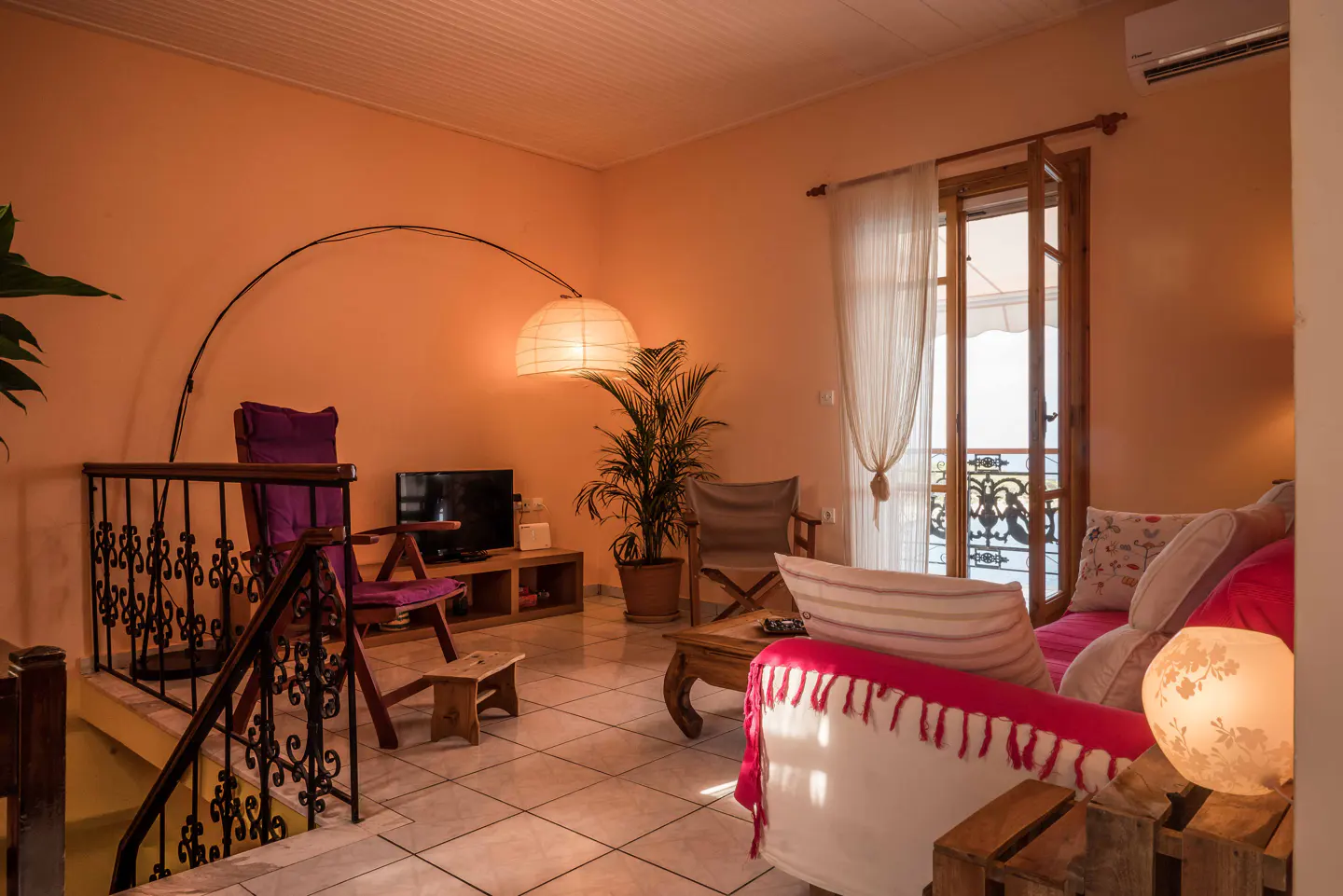 Warm living room with peach walls, tile floor, and balcony view. A purple chair sits near a TV, with a floor lamp arching overhead.