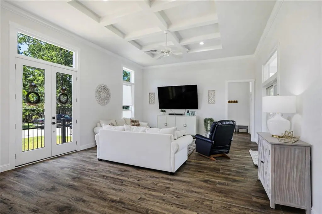 Bright living room with white walls, coffered ceiling, and wood floors. French doors lead to a patio. White sofa, TV, and blue recliner.