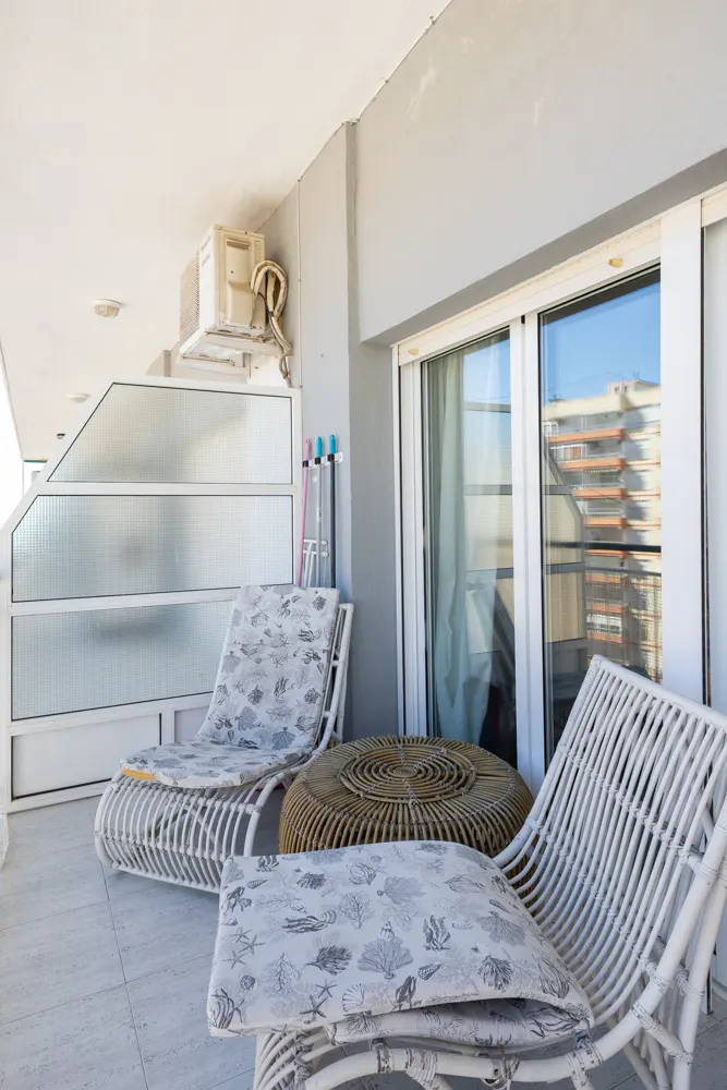 A balcony with white wicker furniture, including two lounge chairs with patterned cushions and a round wicker table. Sliding glass doors reflect a building across the street.