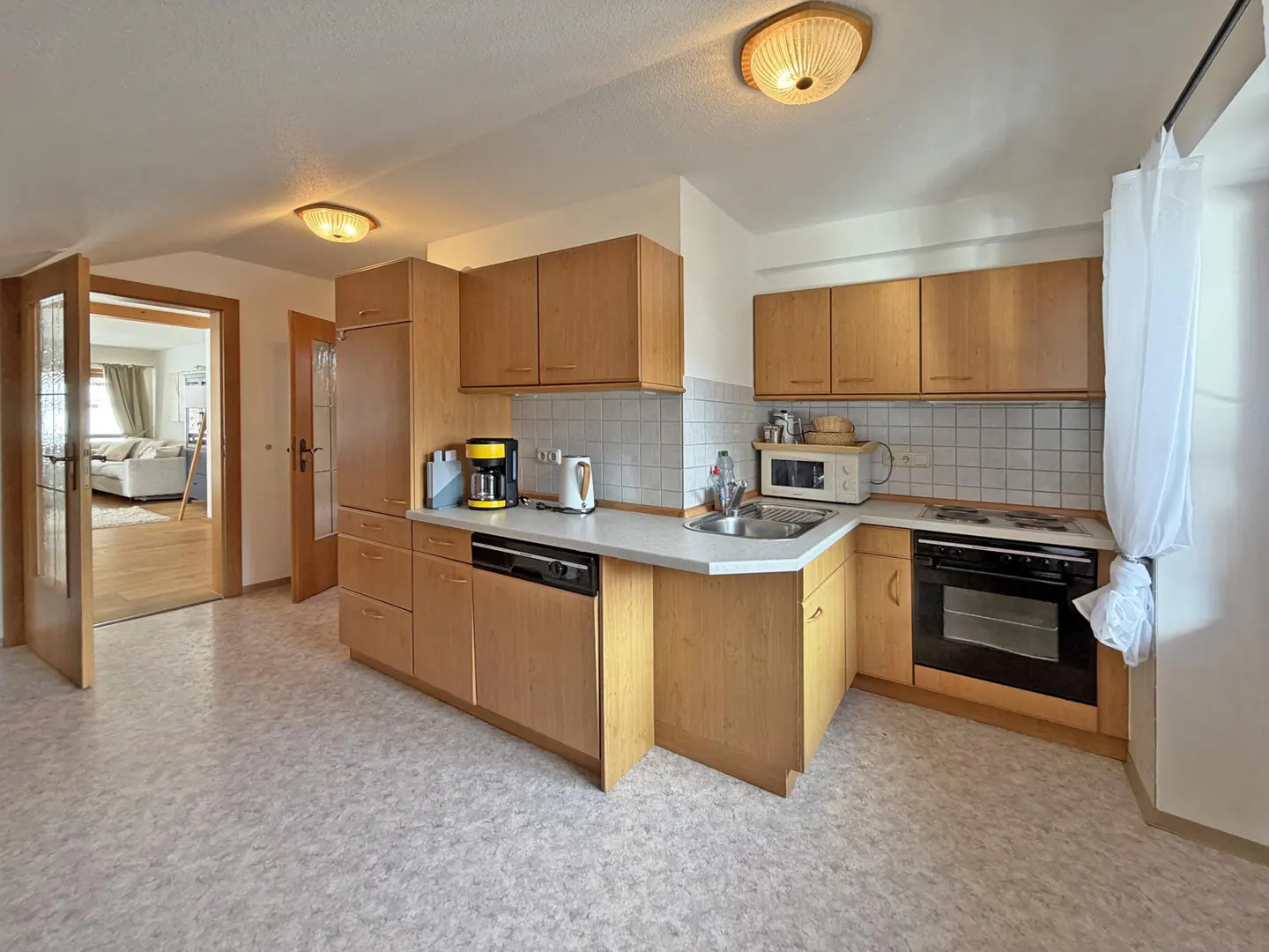 A bright kitchen with light wood cabinets, white countertops, and appliances. A doorway leads to a living room with a sofa.