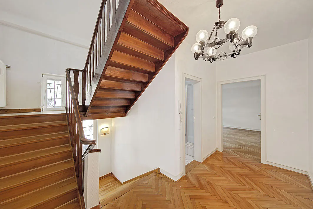 Interior view of a home featuring a wooden staircase, herringbone floors, and a chandelier. White walls and doorways are visible.