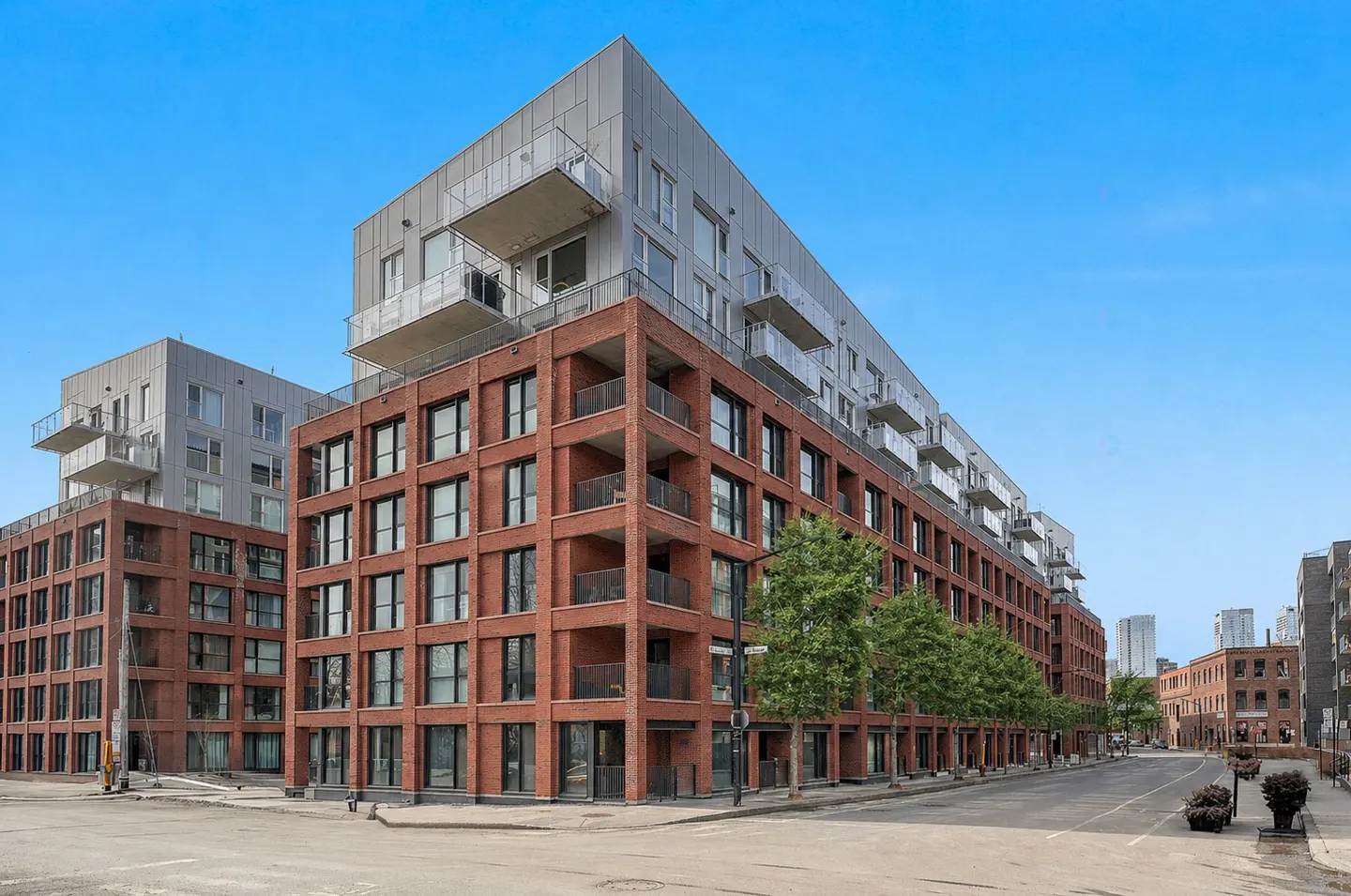 Exterior view of a modern brick apartment building with silver upper floors and balconies under a clear blue sky.