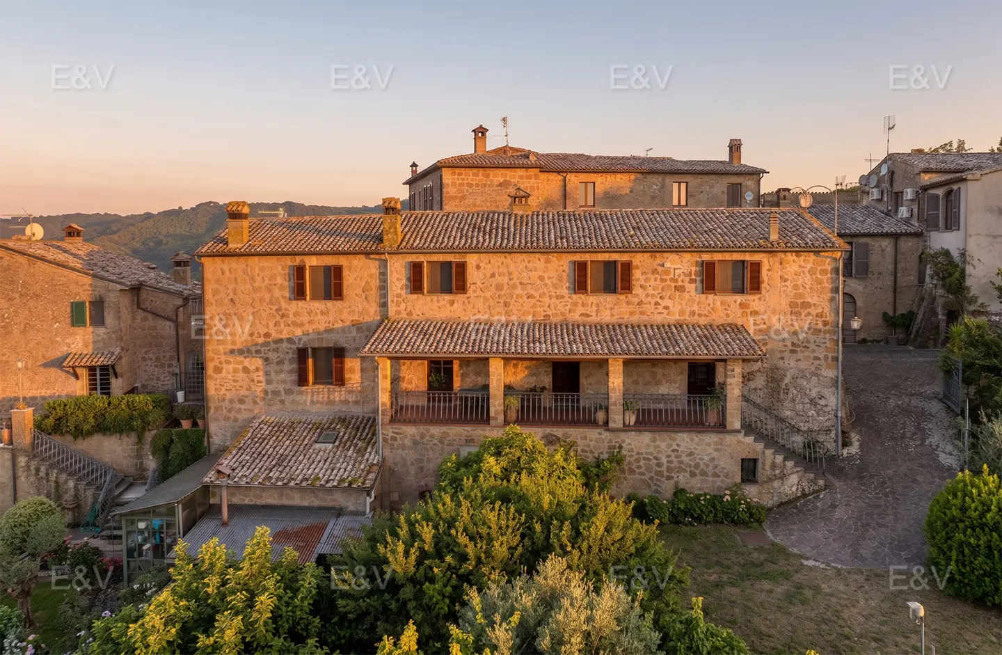 A rustic stone house in Italy at sunset, featuring a covered balcony, tiled roof, and surrounding lush green trees.