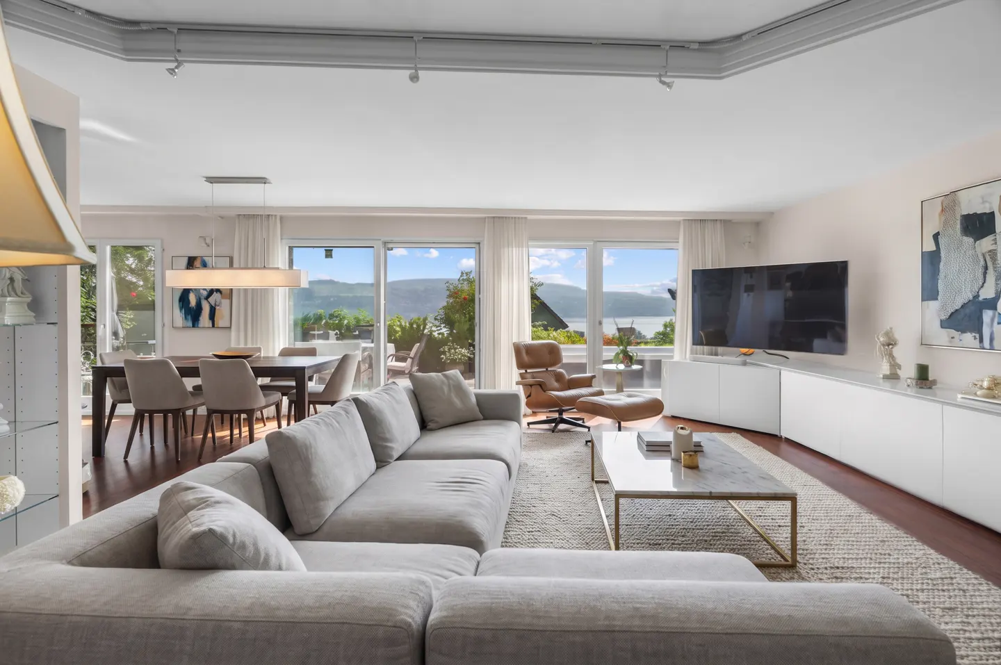 Bright, open living room with a gray sectional sofa, dining table, and a view of a lake through large windows.