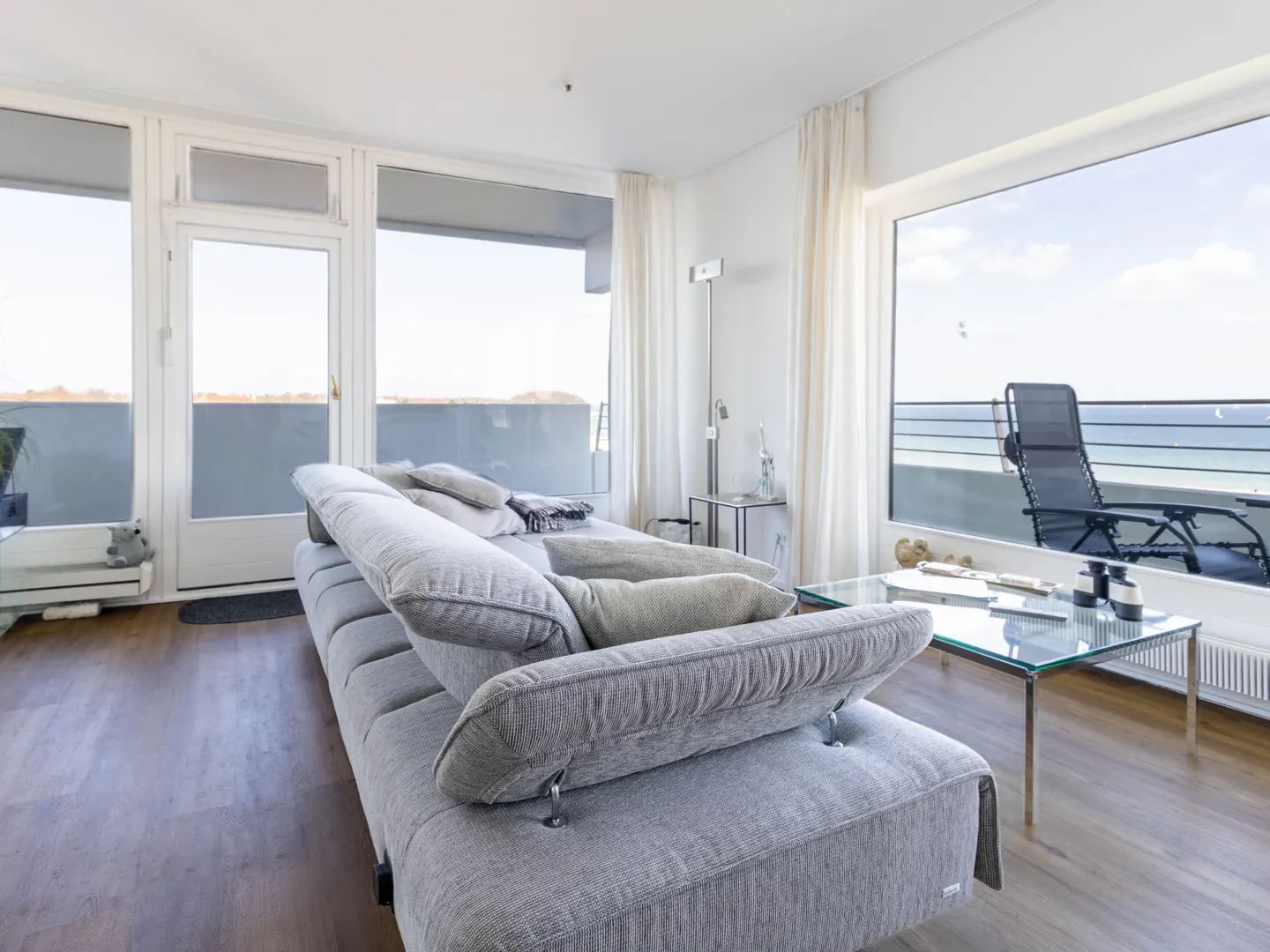 Living room with a gray sofa, glass table, and large windows showing a balcony with a lounge chair and ocean view.
