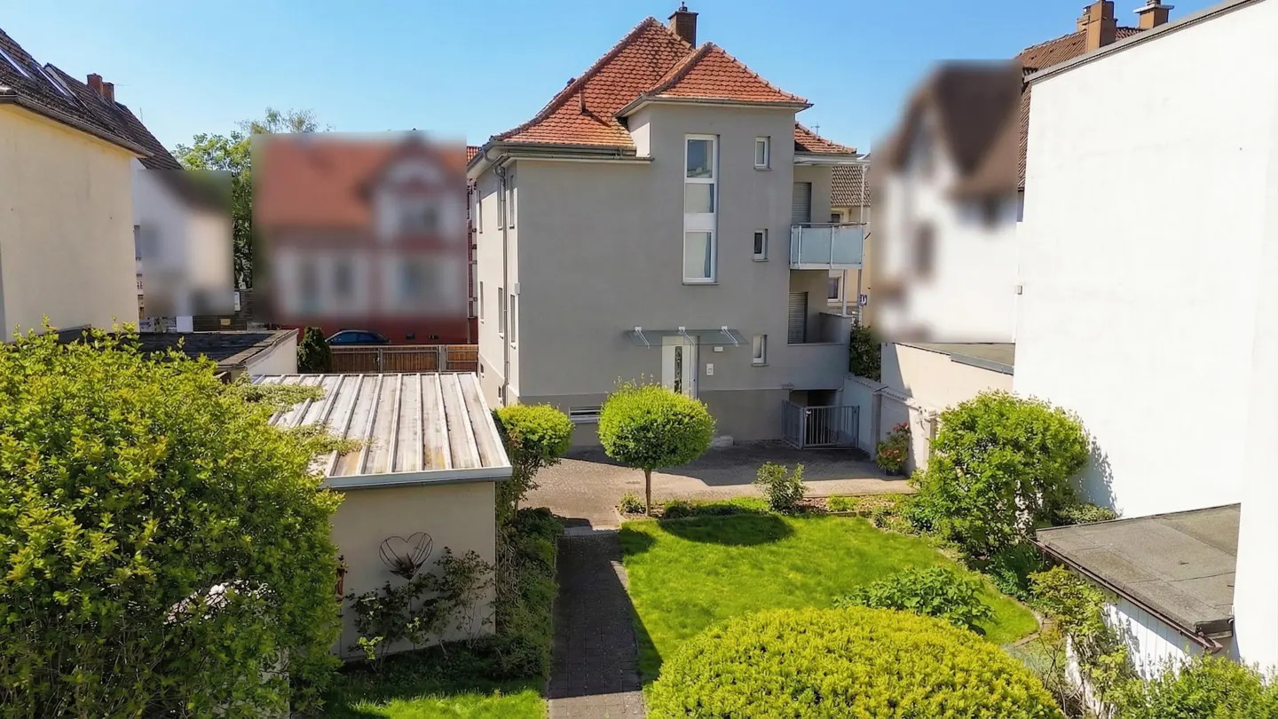 Exterior view of a gray, three-story building with a red tile roof and a small garden with green grass and trees.