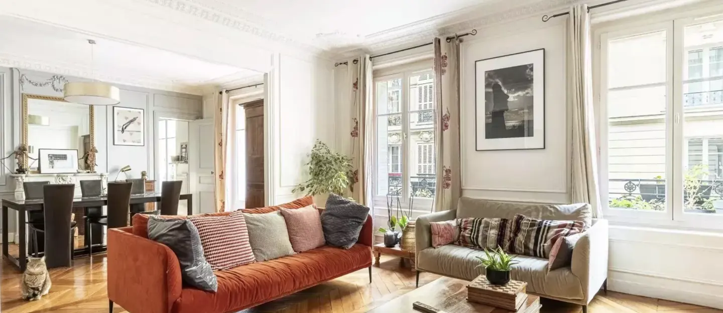 Bright living room with an orange sofa, a beige loveseat, patterned pillows, large windows, and a dining area in the back.