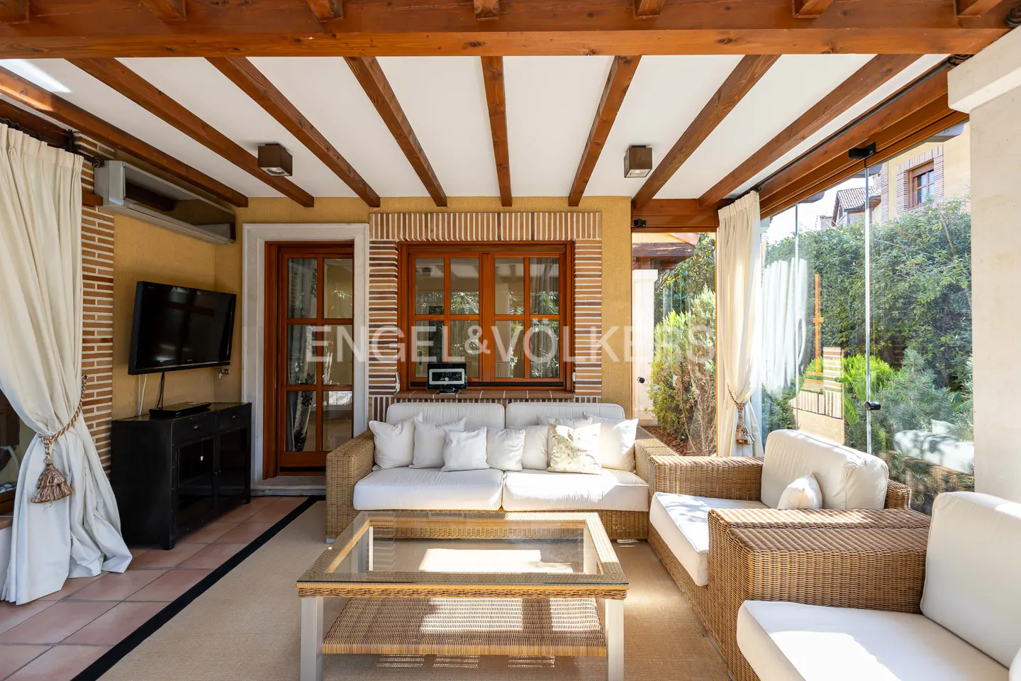 Covered patio with wicker furniture, white cushions, and a glass-topped coffee table. Wood beams line the ceiling. Engel & Volkers logo on window.