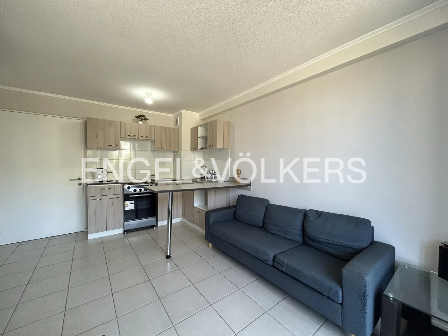 Modern studio apartment with light wood cabinets, a black stove, a breakfast bar, and a dark grey sofa on tiled floors.