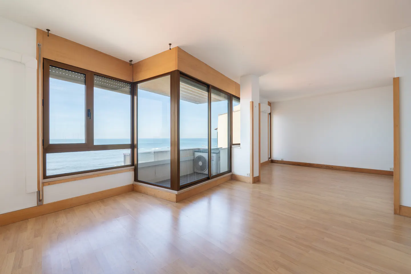 Bright, empty room with light wood floors and white walls. Large windows offer ocean views.