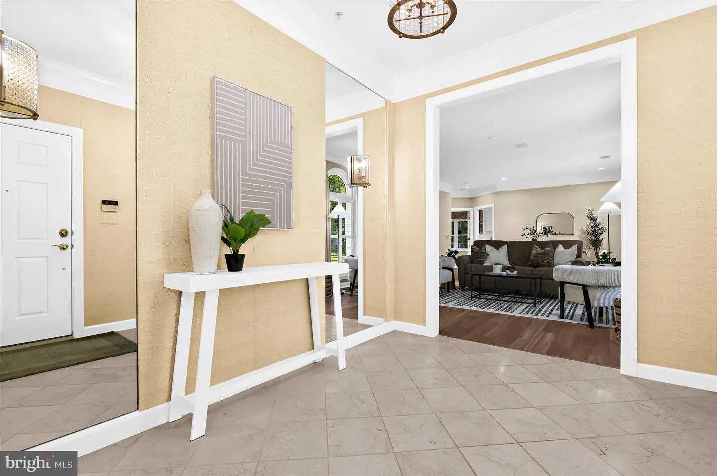 Foyer with beige walls, tile floor, and white trim. A white console table holds a vase and plant. A living room is visible through an archway.