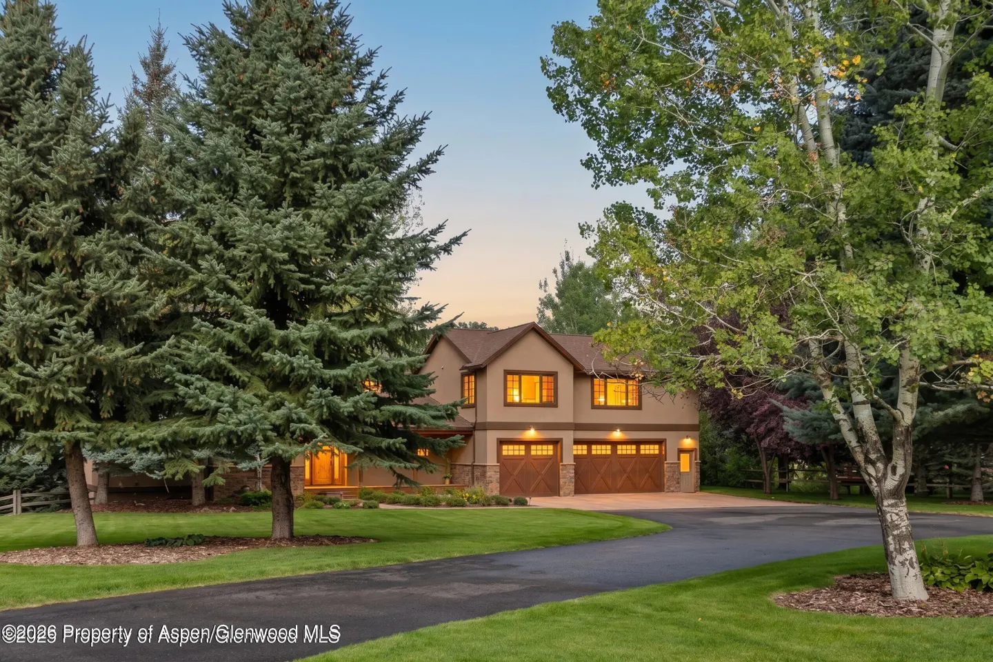 Two-story tan house with brown trim and two-car garage, surrounded by green lawn and trees. Windows are lit.