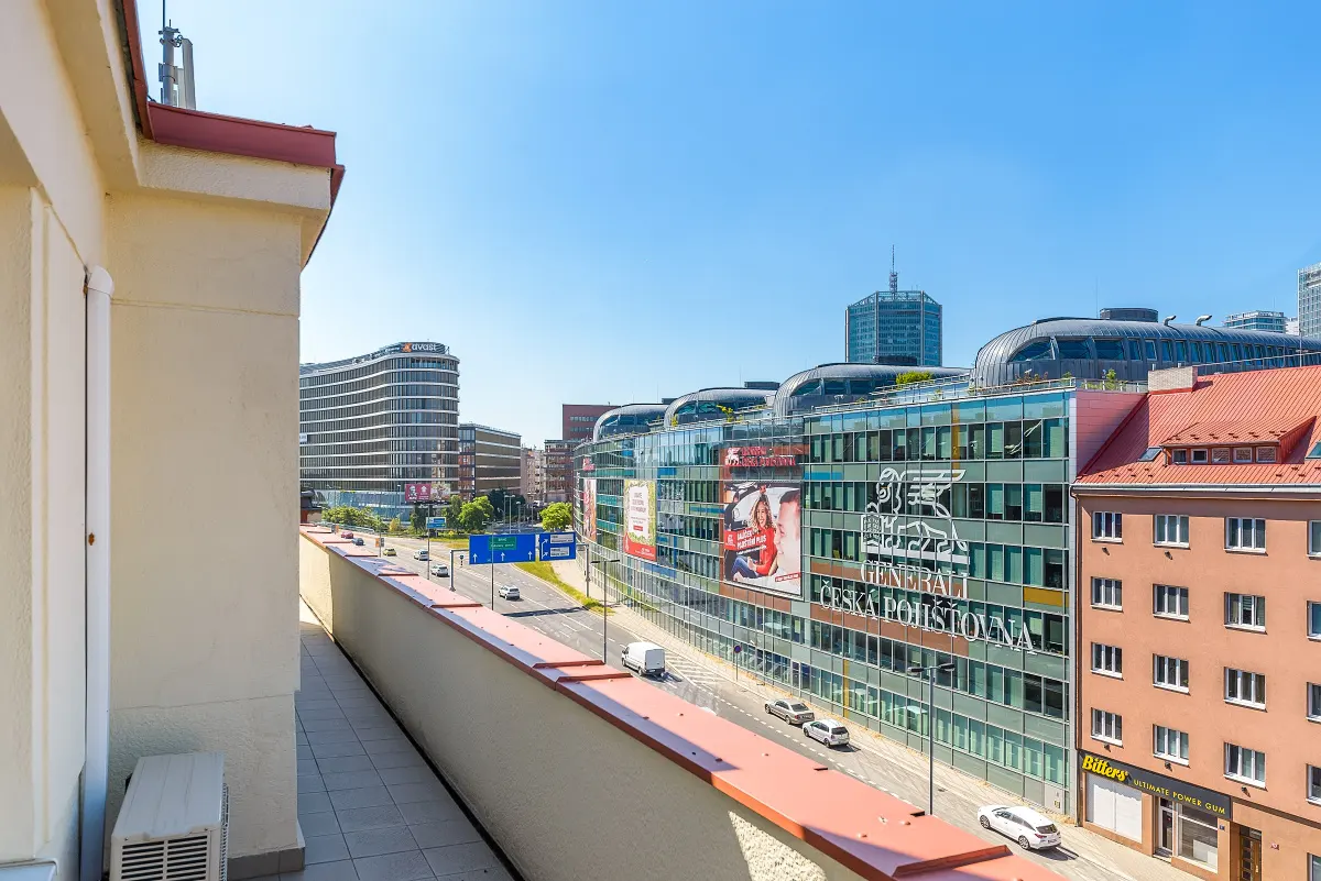 View from a balcony overlooking a sunny city street with modern office buildings, a red-roofed apartment, and blue sky.