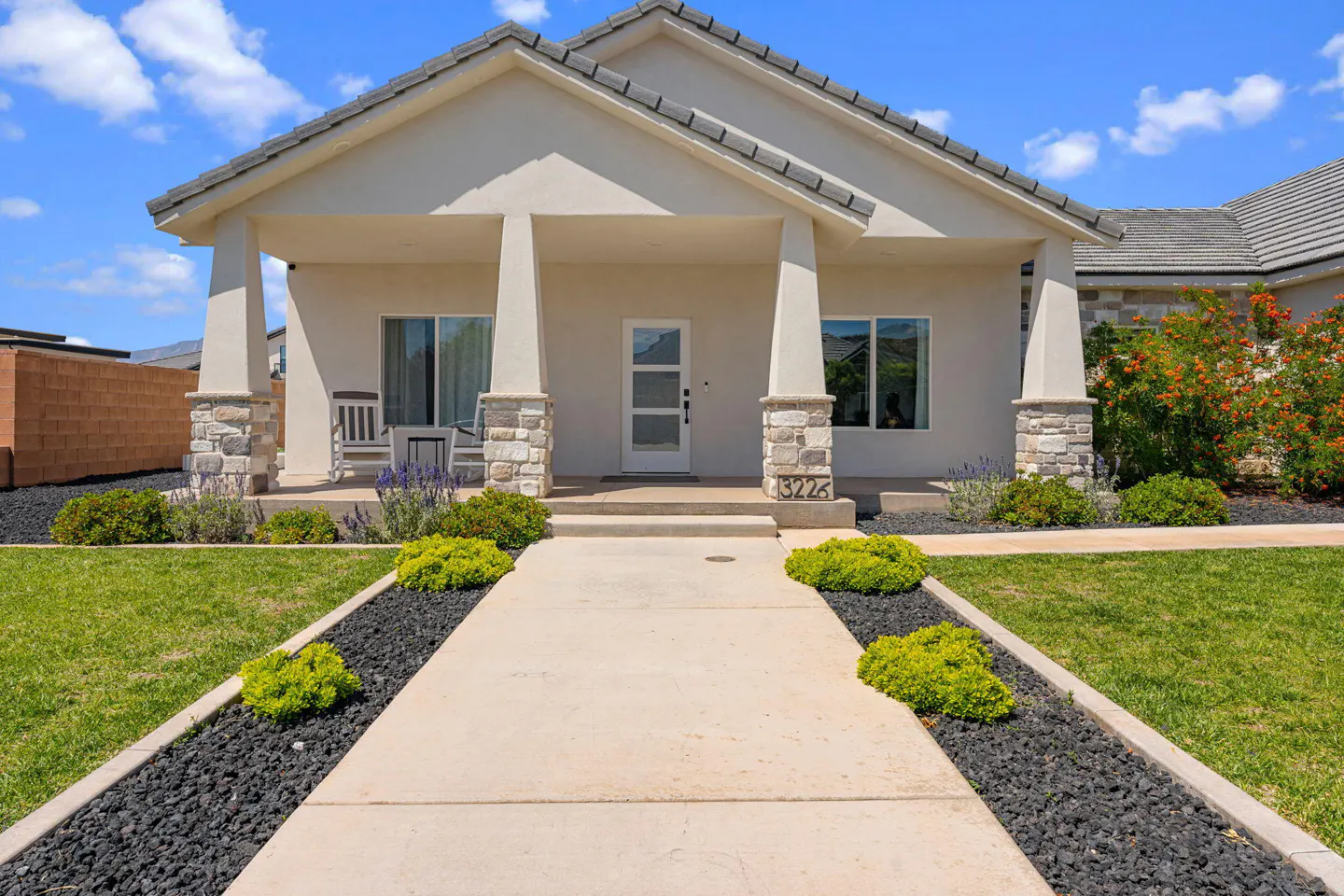 Beige single-story home with a covered porch, stone pillars, and a white front door. A concrete walkway leads to the house, flanked by black rock beds and green bushes.