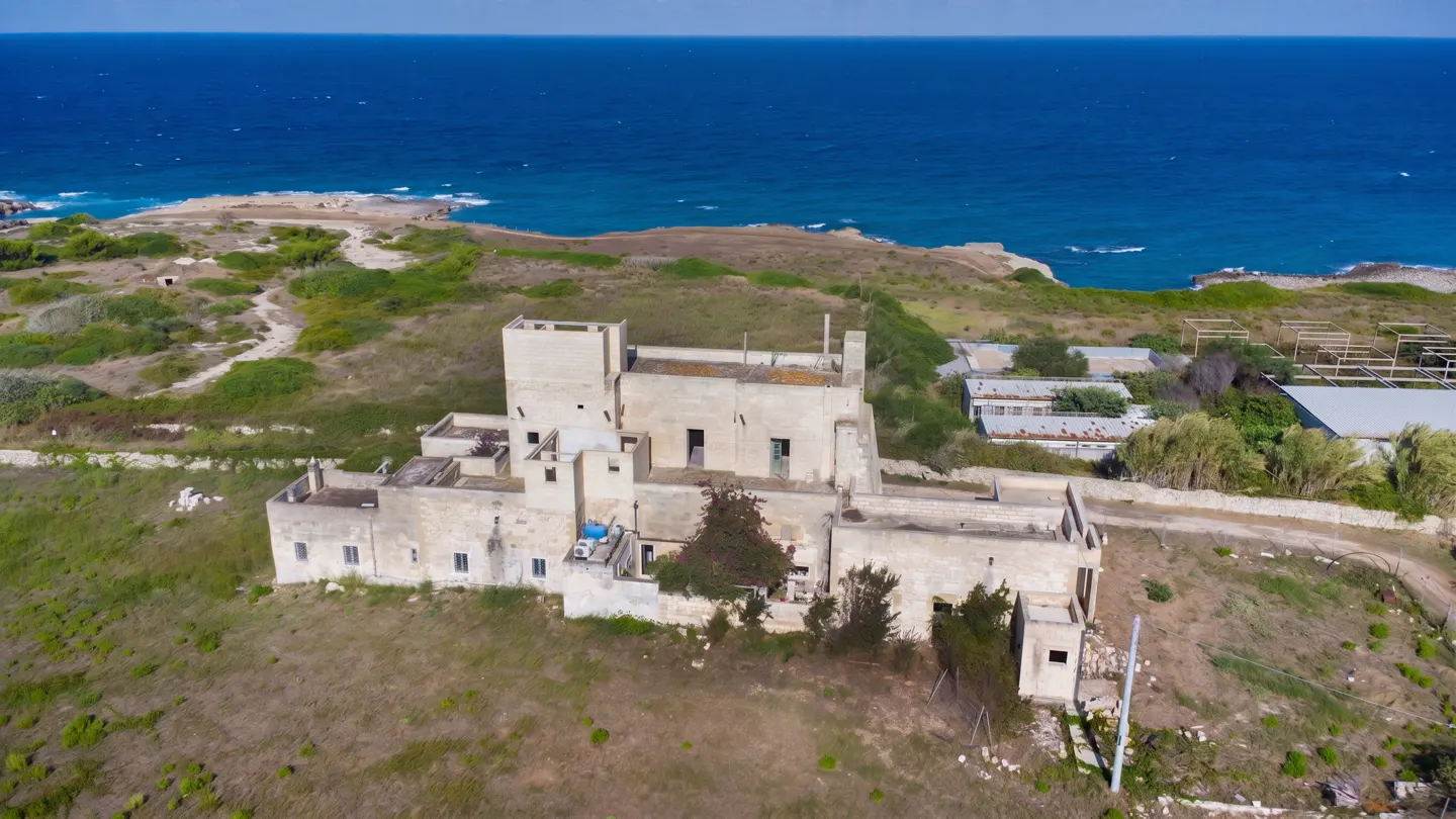 Aerial view of a beige, multi-level building with a flat roof, near the ocean on a sunny day.