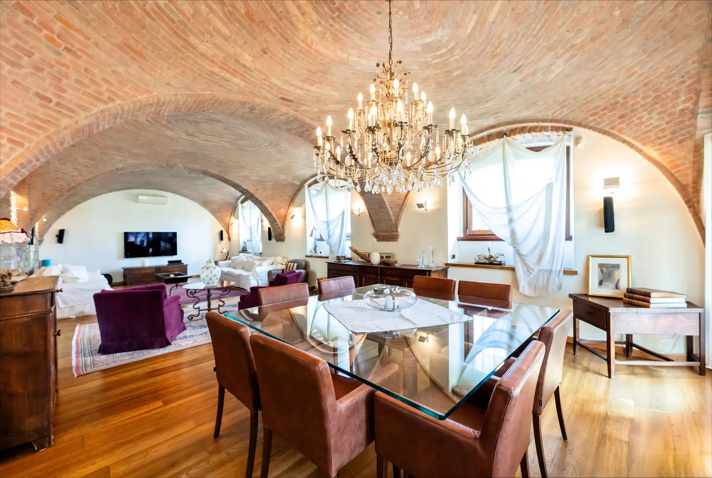 Open-concept living and dining room with brick arched ceiling and chandelier. Glass table with brown leather chairs. Hardwood floors.